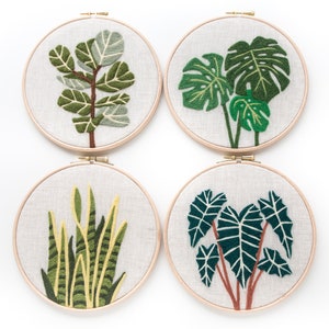 May include: Four embroidery hoops with different plant designs. The hoops are made of wood and have a white fabric background. The plants are embroidered in green, yellow, and brown thread. The designs include a monstera plant, a fiddle leaf fig, a snake plant, and a taro plant.