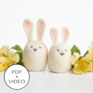 Könnte beinhalten: Zwei weiße Filz-Hasenfiguren mit rosa Ohren und grauen Gesichtern. Die Hasen sitzen auf einer weißen Oberfläche mit gelben Blumen im Hintergrund. Der Text "PDF + VIDEO" befindet sich in einem schwarzen Kreis im Vordergrund.