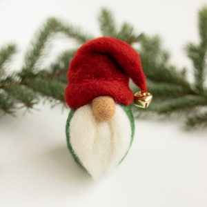 May include: A felt gnome ornament with a red hat, white beard, and green body. The gnome is wearing a gold bell on its hat.