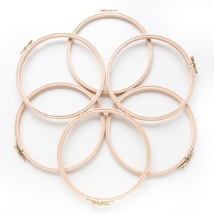 May include: Six wooden embroidery hoops of varying sizes are arranged in a circular pattern on a white background. The hoops are made of natural wood and have a smooth finish.