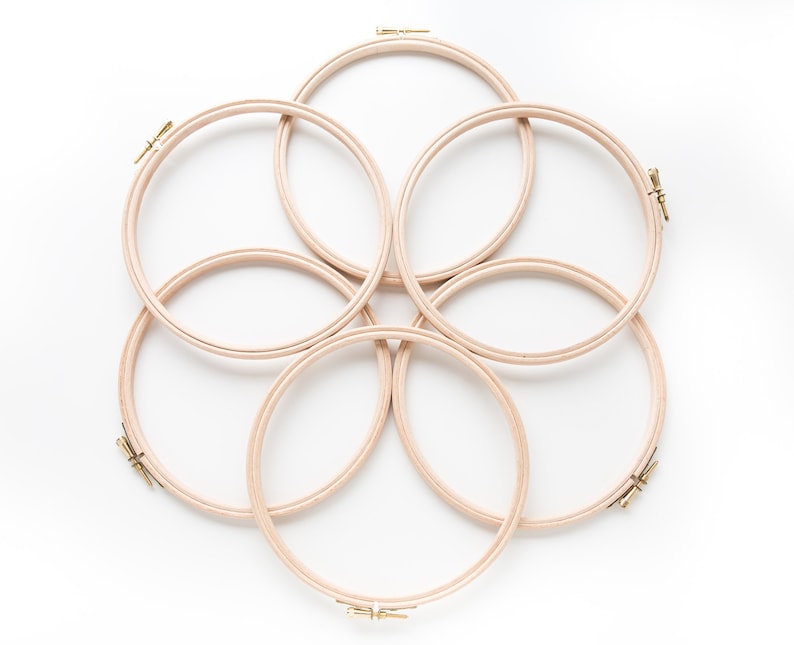 May include: Six wooden embroidery hoops of varying sizes are arranged in a circular pattern on a white background. The hoops are made of natural wood and have a smooth finish.