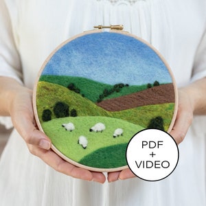 Op de afbeelding: Een ronde borduurring met een viltlandschapsscene. De scène toont glooiende groene heuvels, een bruin veld en een blauwe lucht. Er zijn witte schapen in het groene veld. De ring heeft een wit label met de tekst "PDF + VIDEO".
