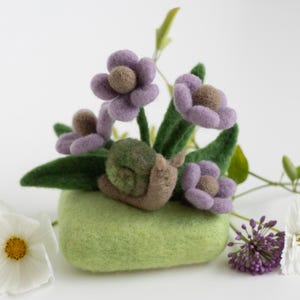 Könnte beinhalten: Eine Filzschnecken-Skulptur mit grüner Schale und braunem Körper, eingebettet in ein Bett aus grünem Filz mit violetten Filzblumen.