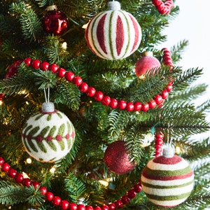 Op de afbeelding: Een kerstboom versierd met rode en witte gestreepte vilten kerstballen, groene vilten kerstballen met bladmotieven en een rode kralenkrans.