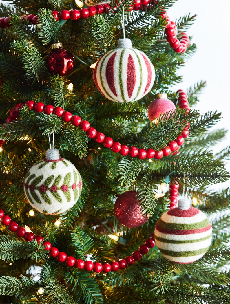Op de afbeelding: Een kerstboom versierd met rode en witte gestreepte vilten kerstballen, groene vilten kerstballen met bladmotieven en een rode kralenkrans.