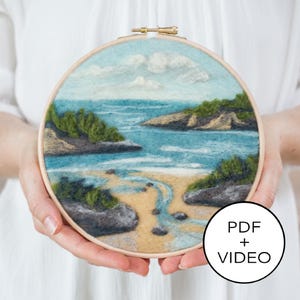 Op de afbeelding: Een close-up van een naaldviltwerk dat een zeelandschap voorstelt met een zandstrand, blauw water en groen gebladerte. Het kunstwerk is ingelijst in een houten hoepel en wordt vastgehouden door handen. De tekst "PDF + VIDEO" is zichtbaar in de rechterbenedenhoek van de afbeelding.
