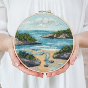 May include: A circular needlepoint embroidery hoop with a felt depiction of a seascape. The scene features a sandy beach with a small stream flowing into the ocean, surrounded by green foliage and rocky outcroppings. The sky is a light blue with fluffy white clouds.