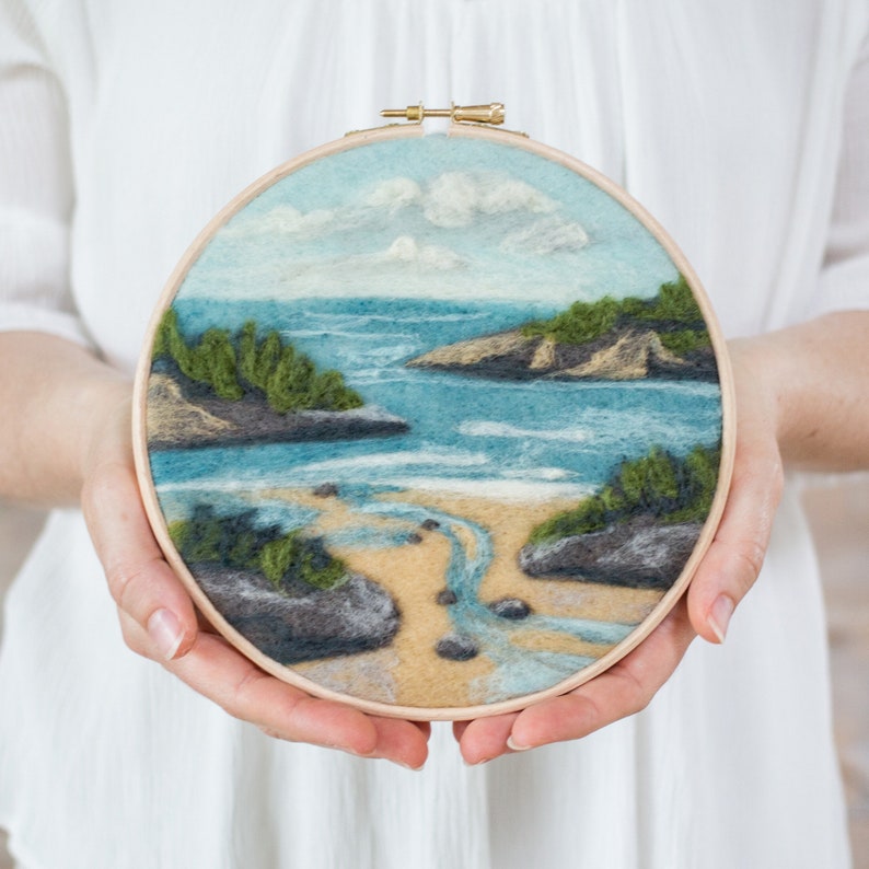 May include: A circular needlepoint embroidery hoop with a felt depiction of a seascape. The scene features a sandy beach with a small stream flowing into the ocean, surrounded by green foliage and rocky outcroppings. The sky is a light blue with fluffy white clouds.