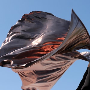 Puede incluir: Una bandera fluida de bronce metálico contra un cielo azul claro. La tela captura la luz, creando reflejos y sombras. La bandera se sostiene en alto, su forma es dinámica y fluida. El efecto general es de movimiento y elegancia.