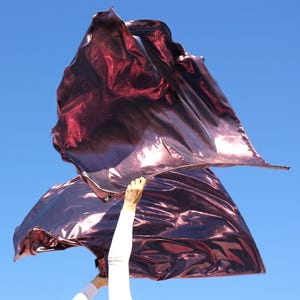 May include: A flowing, burgundy-colored flag against a clear blue sky. The flag has a metallic sheen, catching the light and creating highlights and shadows. The fabric is held aloft by a person wearing a white long-sleeved shirt.