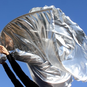 May include: A silver fabric dance flag being held by a person wearing black clothing. The flag is flowing in the wind against a blue sky.
