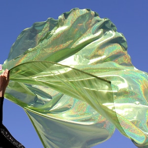 May include: A person holding a large, green, iridescent fabric, or dance flag, in the air. The fabric is flowing in the wind, creating a dramatic effect.