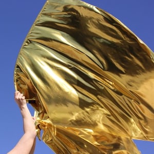 May include: A gold metallic fabric flag being held by a person's hand. The flag is flowing in the wind and has a shiny, reflective surface.