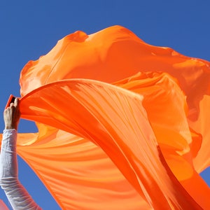 May include: A person holding a bright orange fabric, flowing in the wind against a clear blue sky.