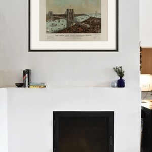 May include: Framed vintage print of the Great East River Suspension Bridge, with boats and buildings. The print is in a black frame, mounted on a white wall above a fireplace. A small blue vase and books are on the mantel.