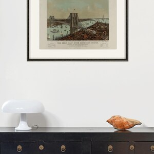 May include: Framed vintage print of the Great East River Suspension Bridge, with boats and buildings. The print is in a black frame, displayed above a black cabinet with brass hardware. A white mushroom lamp and a seashell are on the cabinet.