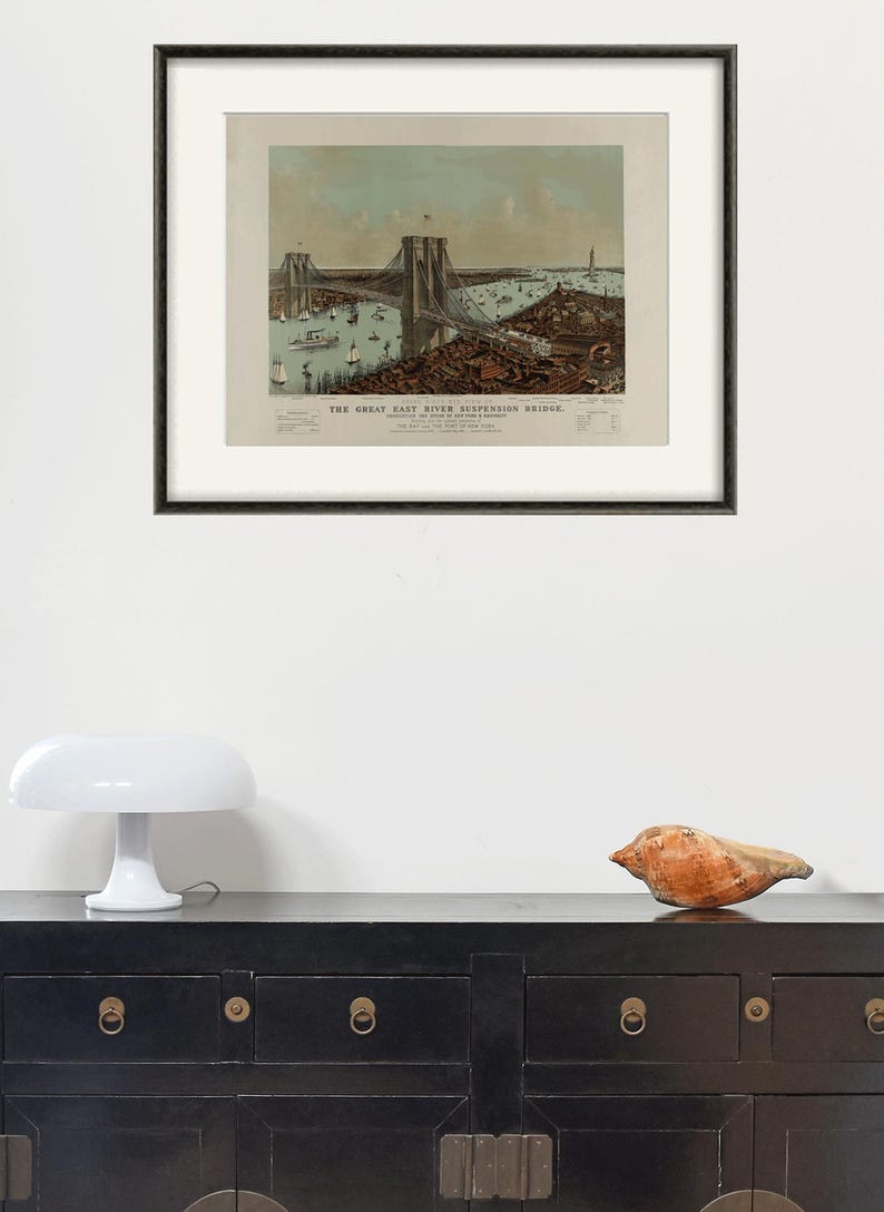 May include: Framed vintage print of the Great East River Suspension Bridge, with boats and buildings. The print is in a black frame, displayed above a black cabinet with brass hardware. A white mushroom lamp and a seashell are on the cabinet.