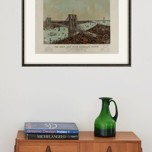 May include: Framed vintage print of the Great East River Suspension Bridge, featuring a detailed illustration of the bridge, boats, and surrounding cityscape. The print is in a black frame and is displayed above a wooden cabinet. A green glass pitcher and books are on the cabinet.