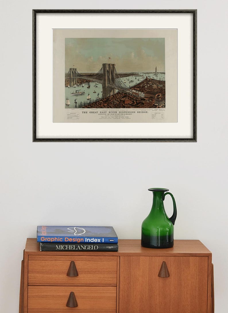May include: Framed vintage print of the Great East River Suspension Bridge, featuring a detailed illustration of the bridge, boats, and surrounding cityscape. The print is in a black frame and is displayed above a wooden cabinet. A green glass pitcher and books are on the cabinet.