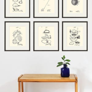 Puede incluir: Seis dibujos de patentes en blanco y negro enmarcados de equipo de golf. Los dibujos incluyen un palo de golf, una pelota de golf, un zapato de golf y un tee de golf. Los dibujos se muestran en una pared blanca sobre una mesa de madera.