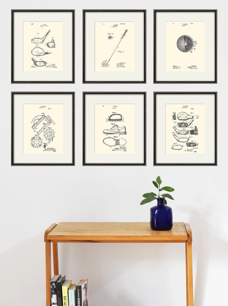 Puede incluir: Seis dibujos de patentes en blanco y negro enmarcados de equipo de golf. Los dibujos incluyen un palo de golf, una pelota de golf, un zapato de golf y un tee de golf. Los dibujos se muestran en una pared blanca sobre una mesa de madera.