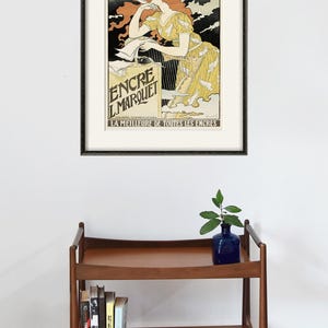 May include: A framed vintage poster advertising "Encre L.Marquet" ink. The poster features a woman with red hair writing with a quill pen. The text on the poster reads "La Meilleure de Toutes les Encres" which translates to "The Best of All Inks".