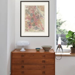 May include: A vintage map of Vienna, Austria, framed in black, hangs on a white wall above a wooden dresser with three drawers. A white lamp sits on the dresser, and a small blue bowl and a larger gray bowl are on the dresser. A silver rocket-shaped lava lamp sits on a windowsill.