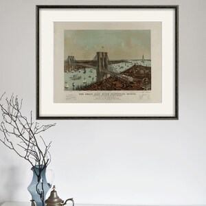 May include: Framed print of the Great East River Suspension Bridge, featuring a detailed illustration of the bridge, boats, and buildings. The print is in a black frame with a white mat. A decorative vase and teapot are in the foreground.