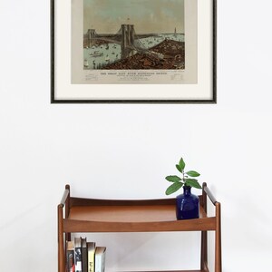 May include: Framed vintage print of the Great East River Suspension Bridge, with a black frame and off-white matting. The print depicts the bridge, boats, and buildings. A wooden shelf with books, a blue vase, and a decorative bowl is below.