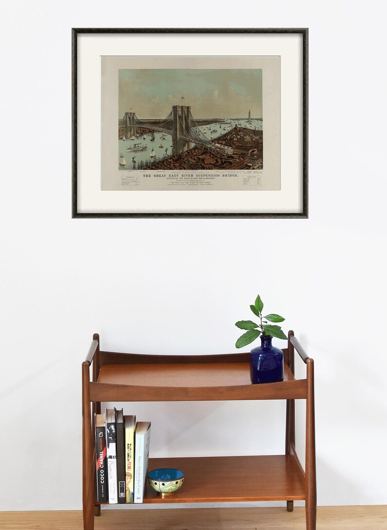 May include: Framed vintage print of the Great East River Suspension Bridge, with a black frame and off-white matting. The print depicts the bridge, boats, and buildings. A wooden shelf with books, a blue vase, and a decorative bowl is below.