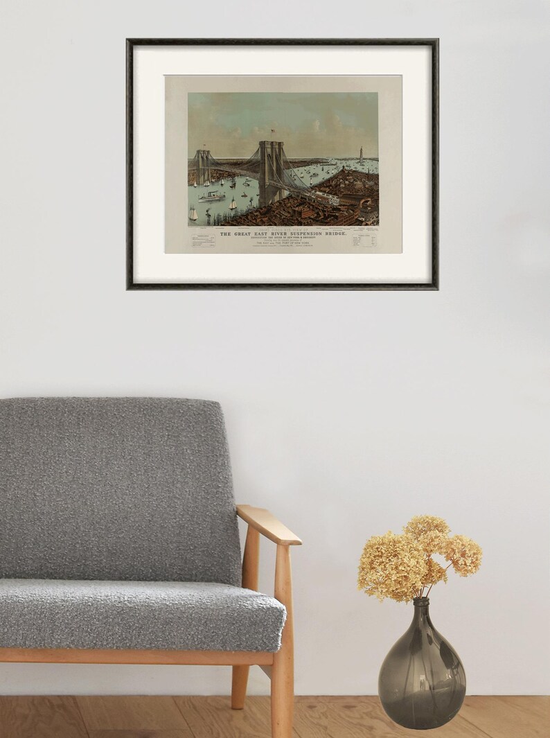 May include: Framed vintage print of the Great East River Suspension Bridge, featuring a detailed illustration of the bridge, boats, and surrounding cityscape. The print is in a black frame and is displayed on a white wall.
