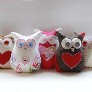 May include: A collection of handmade owl-shaped plush toys. Each owl has button eyes, a heart-shaped appliqué, and unique fabric patterns in various colors, including white, brown, pink, and gray. The owls are arranged in a row.