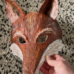 May include: A fox mask with a textured, hand-painted finish in shades of reddish-brown, orange, and white. The mask features pointed ears, almond-shaped eye openings, and a black nose, showcasing visible brushstrokes across the surface.