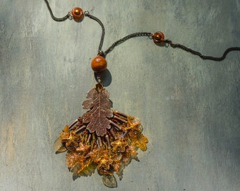 May include: A handcrafted necklace featuring a bronze-coloured oak leaf pendant with intricate detailing. The pendant is adorned with amber-coloured beads and delicate floral accents. The necklace has a dark chain with amber-coloured beads.