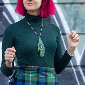 May include: A woman with pink hair wearing a dark green turtleneck sweater and a plaid kilt. She is wearing a silver chain necklace with a green and silver pendant.