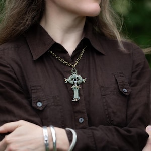May include: A brown button-down shirt with a unique pendant necklace. The necklace has a detailed, ornate design with a chain. Silver bracelets adorn the wrist. The overall aesthetic is earthy and bohemian.
