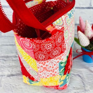 May include: A small red and yellow patchwork fabric zippered pouch with a red fabric handle. The pouch is made of a variety of fabrics with different patterns, including polka dots, flowers, and geometric shapes.