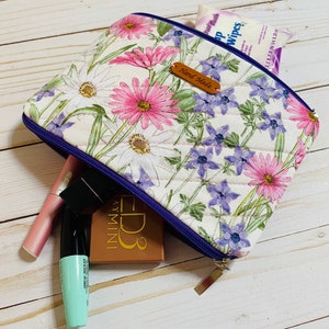May include: A white quilted zippered pouch with a floral print featuring pink, purple, and white flowers. The pouch has a blue zipper and a brown leather tag that says "Hand Made". The pouch is open and contains makeup items, including a pink lipstick, a blue mascara, and a brown eyeshadow palette.