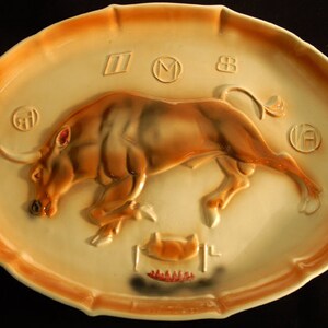 May include: A ceramic oval platter with a raised image of a brown bull charging. The platter has a cream-colored background with brown accents and the numbers "1908" in the center.