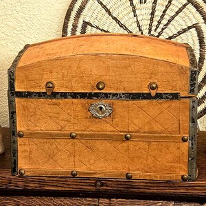 May include: A vintage-style trunk with a domed top and a light brown exterior. The trunk features metal accents, including corner protectors, a keyhole, and decorative studs. The trunk is made of a textured material with a diamond pattern.