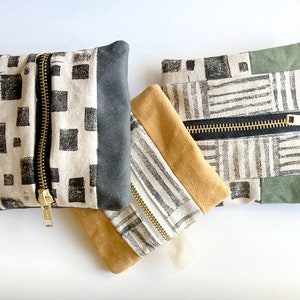 May include: Three small zippered pouches with different fabric patterns. The pouches are made of a white fabric with black geometric designs. The pouches are stacked on top of each other. The top pouch is green, the middle pouch is brown, and the bottom pouch is black.