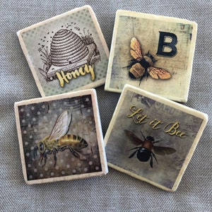 May include: Four square ceramic coasters with bee and honey themed designs. The coasters feature a beehive, bees, and the words "Honey", "B", and "Let it Bee".