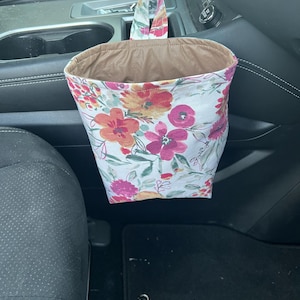 May include: A floral print car trash bag with a brown interior. The bag is white with a pattern of pink, orange, and yellow flowers. It hangs from the gear shift.