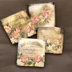 May include: Four square ceramic coasters with floral designs and vintage text. The coasters feature pink and white flowers, green leaves, and handwritten text in French and English. The text includes "Blanc de Perles Liquide", "la Ville & le Theatre", "Prepare Par", "Carte Post", and "Paris".