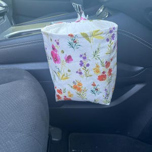 May include: A fabric car trash bag featuring a white background with a colorful floral print of red, pink, purple, and yellow wildflowers. The bag is designed to hang from the gear shift lever of a vehicle, providing a portable waste container.