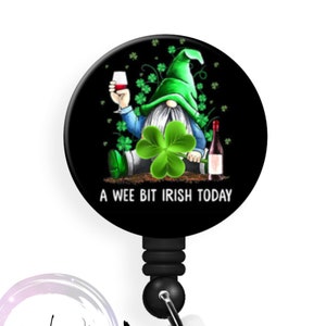 May include: A black and green badge reel with a cartoon gnome holding a glass of wine and a bottle of wine. The gnome is wearing a green hat and sitting on a bed of green shamrocks. The text "A wee bit Irish today" is printed on the reel.