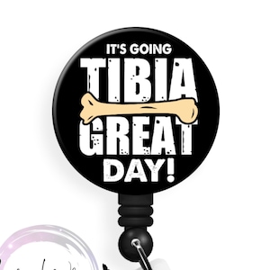 May include: A black badge reel with the text "IT'S GOING TIBIA GREAT DAY!" and a bone graphic. The reel has a retractable cord and a clear plastic strap with a snap closure. The Badge Whimsy logo is in the lower left corner.