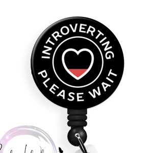 May include: Black and white badge reel with a black circle that says "Introverting Please Wait" in white text. A red heart is in the center of the circle.