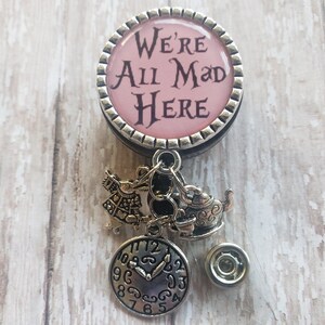 May include: A silver and pink badge holder with a charm of the Mad Hatter, a teapot, and a clock. The badge holder has a pink circle with the text "We're All Mad Here".