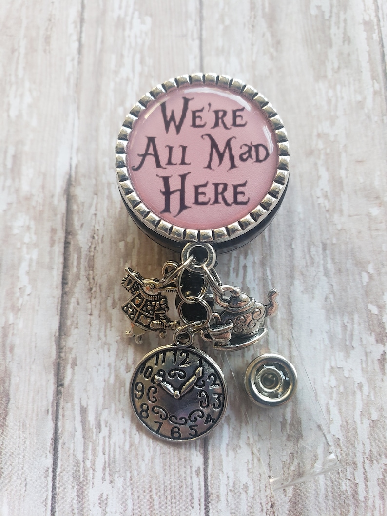 May include: A silver and pink badge holder with a charm of the Mad Hatter, a teapot, and a clock. The badge holder has a pink circle with the text "We're All Mad Here".